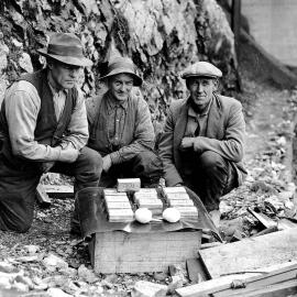 Three miners displaying twelve gold ingots on an upturned crate at the Blackwater Mine in Waiuta. 1932.