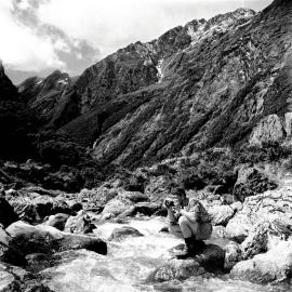 A man taking a photograph at an unidentified river on the track between Blue River and Māori Saddle near Haast Pass.ca.1940- 1959.