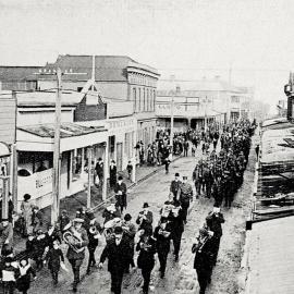 A procession along Revell Street headed by the Municipal band before the unveiling of a memorial to Westland pioneers, Hokitika.1914.