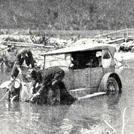 Two men securing a rope to a car, which is in shallow water, on the road to Punakaiki.1924.