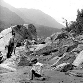 Three people on a sandy riverbank with boulders, Westland`.ca.1890`s-1910`s.