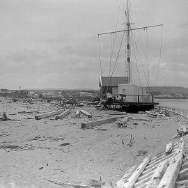 Two small buildings on the Hokitika breakwater, with the larger settlement of Hokitika visible in the background.ca.1900-1916.